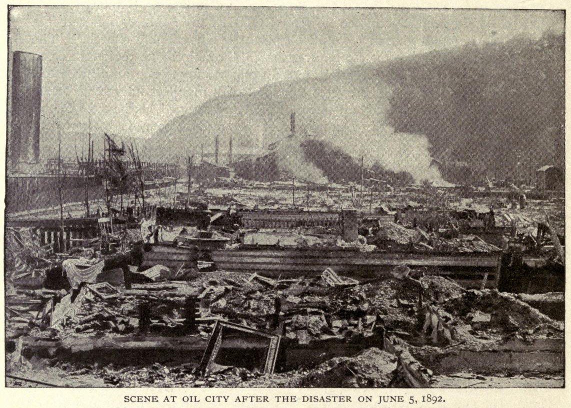 Schwarzes und weißes Foto einer Stadt nach einer Katastrophe am 5. Juni 1892, das Gebäude, Pfähle, Rauch, einen Hügel im Hintergrund und Text unten zeigt.