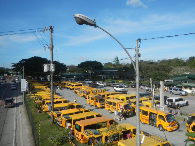 Eine Reihe gelber Schulbusse, die neben einer Straße mit Fußgängern auf dem Gehweg, Strommasten, Bäumen, Gebäuden und einem bewölkten Himmel im Hintergrund geparkt sind.