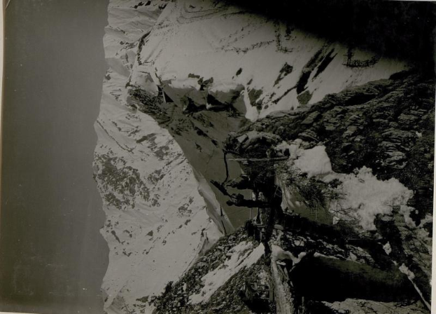 Schwarzes und weißes Foto eines Mannes in Kletterausrüstung, der einen schneebedeckten Berg hinaufsteigt, mit dem Gipfel des Mount Everest im Hintergrund.