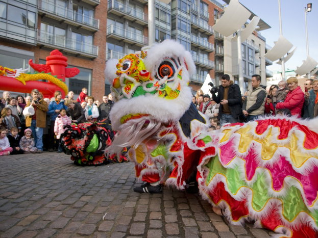 Ein lebendiges chinesisches Neujahrsfest in Amsterdam mit einem Löwen tanzen vor einem Publikum, einige halten Kameras, vor einem Hintergrund von Gebäuden, Laternenmasten und einem klaren blauen Himmel.