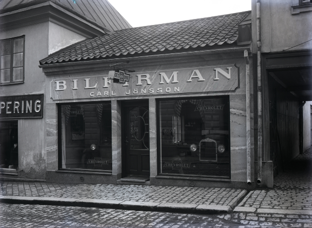Ein Schwarz-Weiß-Foto eines Gebäudes mit einer Tafel, die "Bilferman" lautet, mit Fenstern, einer Tür und Tafeln mit Text, sowie einer Straße am unteren Bildrand.