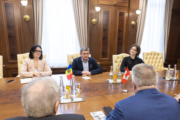 Eine Gruppe von Menschen sitzt um einen Holztisch in einer förmlichen Umgebung und diskutiert mit verschiedenen Gegenständen, darunter Flaggen und Dokumente, was ein Thema im Zusammenhang mit der Europäischen Union nahelegt.