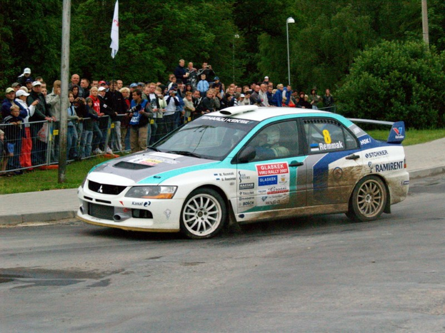 Ein Rallyeauto mit Texten und Logos fährt an einer Zuschauermenge vorbei, die Kameras hält, mit Geländern, Fahnenmasten und Bäumen im Hintergrund.