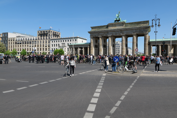 Eine große Gruppe von Menschen, die auf Fahrrädern eine Straße entlang des Brandenburger Tors in Berlin, Deutschland, fährt, mit Bäumen, Gebäuden, Laternenmasten und Verkehrszeichen an der Straße und einem Bogen mit Säulen und einer Statue oben drauf im Hintergrund.