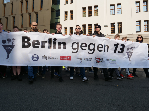 Eine Gruppe von Menschen geht mit einem Banner, auf dem "Berlin gegen 13" steht, die Straße entlang, mit Gebäuden und Himmel im Hintergrund.