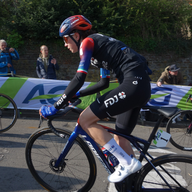 Professionelle Radrennfahrerin Katie Archibald fährt ein Fahrrad die Straße hinunter, trägt einen Helm, eine Schutzbrille, Handschuhe und Schuhe und hat eine Menge und ein Banner im Hintergrund.