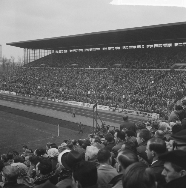 Ein Schwarz-Weiß-Foto eines vollen Stadions mit Menschen, die ein Fußballspiel anschauen, mit Bannern, Pfählen, einer Hütte, Bäumen, einem Turm und einem bewölkten Himmel im Hintergrund.