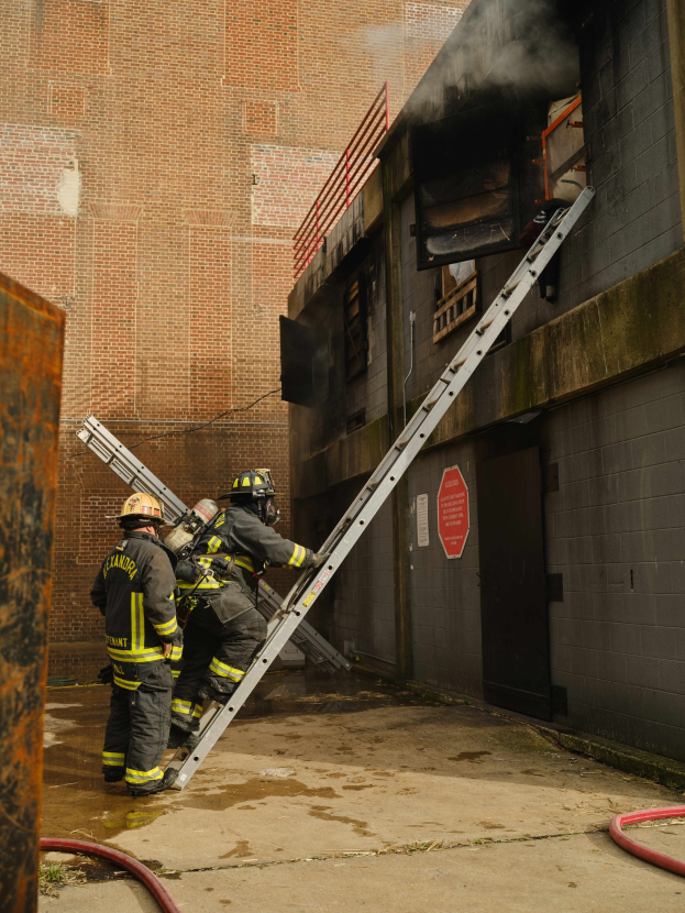 Feuerwehrleute in Helmen klettern eine Leiter zu einem raucherfüllten Gebäude hoch, während ein Metallobjekt und Bodenrohre sichtbar sind.