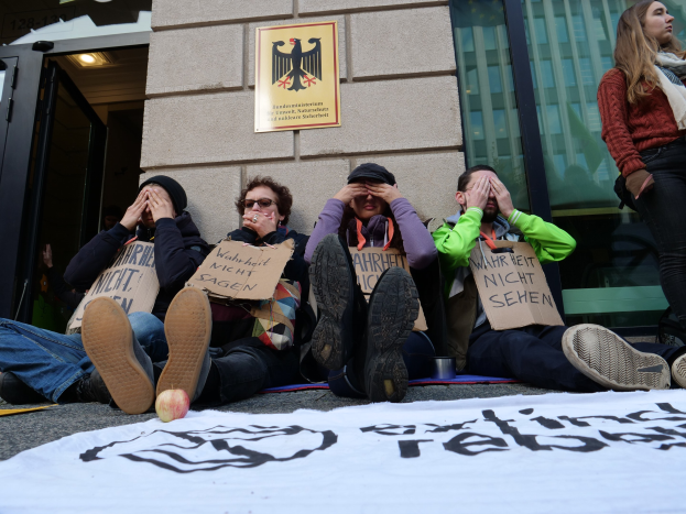 Eine Gruppe von Menschen sitzt vor einem Gebäude auf dem Boden und hält Schilder, mit einem Banner auf der Straße und einer Frau stehend rechts; eine Wand mit einer Tafel und ein Gebäude mit Glasfenstern und einer Tür sind im Hintergrund zu sehen.
