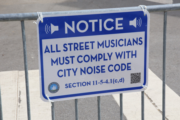 Anschlagtafel an einem Zaun mit einem Schild, auf dem "Hinweis: Alle Straßenmusiker müssen die Stadtlärmvorschriften einhalten" steht und eine Straße im Hintergrund.
