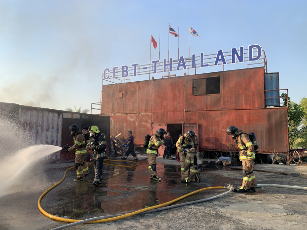 Feuerwehrleute in Helmen sprühen Wasser auf ein Gebäude mit Text, während Fahnen, Bäume und ein blauer Himmel im Hintergrund zu sehen sind.