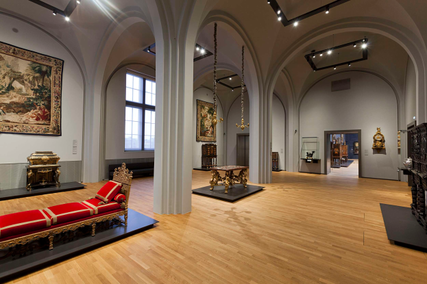Ein geräumiger Raum im Kunsthistorischen Museum in Wien, Österreich, gefüllt mit Möbeln, Wandgemälden und dekorativen Elementen wie einem Sofa mit Kissen, Tischen mit verschiedenen Gegenständen, Säulen, Fenstern und Deckenleuchten.