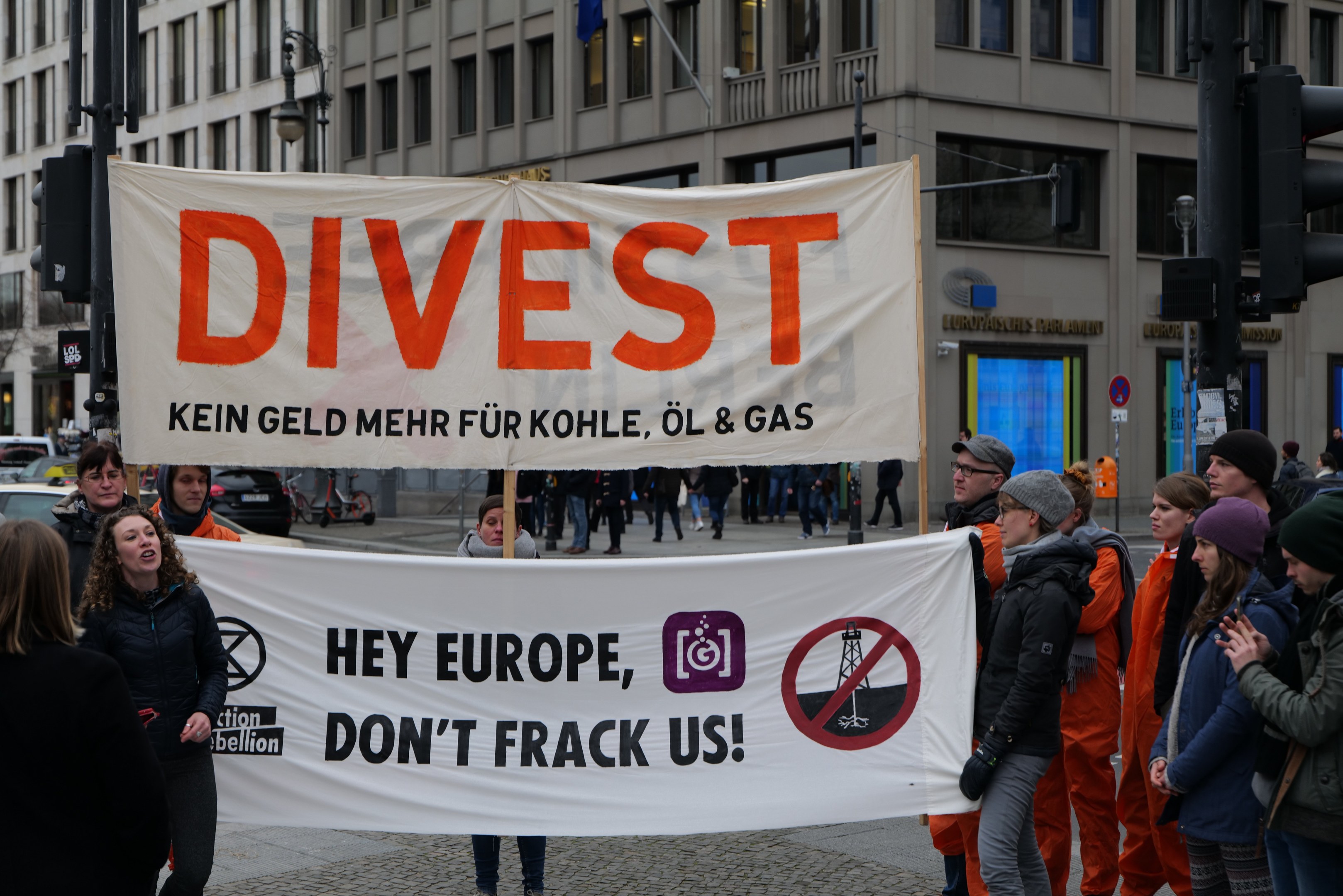 Menschen halten ein "Divest Hey Europe, Don't Frack Us"-Schild auf einer Straße mit Gebäuden, Fahrzeugen und Bäumen im Hintergrund.