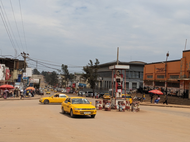 Ein gelbes Taxi fährt auf einer belebten Stadtstraße mit Gebäuden an den Seiten, während Fußgänger mit Regenschirmen auf dem Gehweg unter bewölktem Himmel gehen.