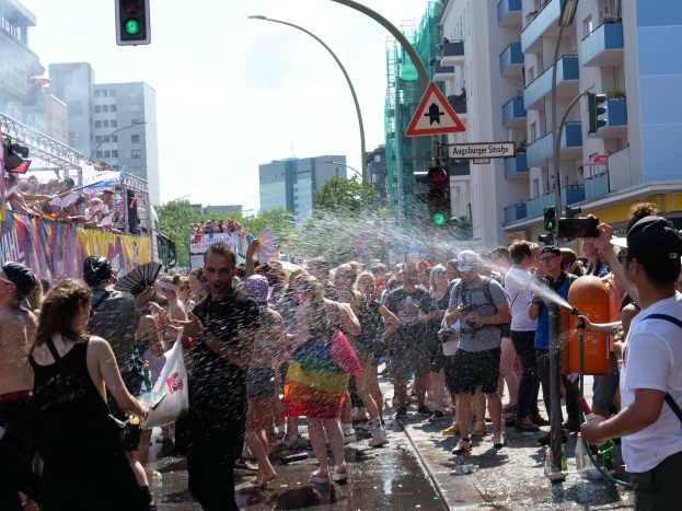 Menschen auf einem Christopher Street Day (CSD) mit Wasser bespritzen sich gegenseitig und halten Gegenstände in den Händen, mit einer Fahne links und Gebäuden, Bäumen und Ampeln im Hintergrund.