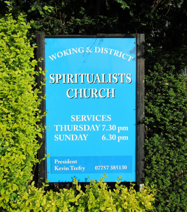 Ein hölzernes Schild mit der Aufschrift "Woking & District Spiritualists Church" steht draußen, umgeben von Grün mit einem Gebäude im Hintergrund.