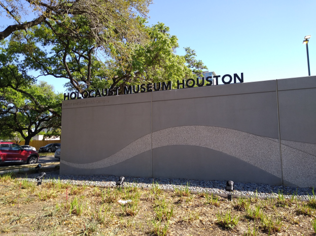 Außenansicht des Holocaust-Museums in Houston mit eingravierter Textwand, verstreuten Steinen, Grünfläche, Bäumen, vorbeifahrenden Fahrzeugen, einem Straßenpfahl und einem bewölkten Himmel.