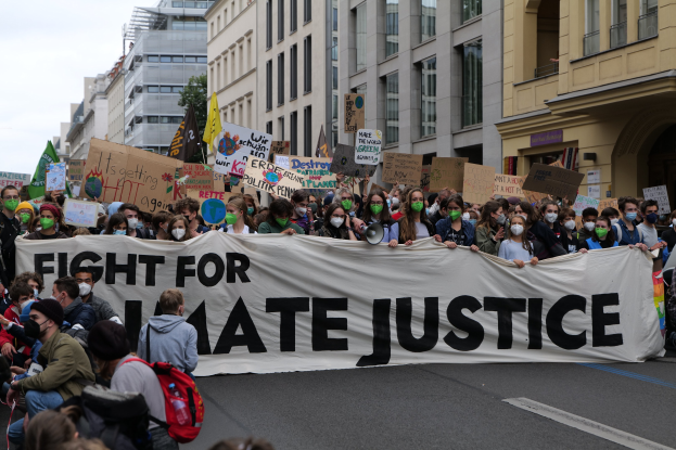 Eine große Gruppe von Menschen marschiert mit einem Banner, auf dem 'Kämpfe für Hassjustiz' steht, durch eine Straße. Einige tragen Masken und halten Schilder oder Taschen, vor dem Hintergrund von Gebäuden, Bäumen und einem klaren blauen Himmel.