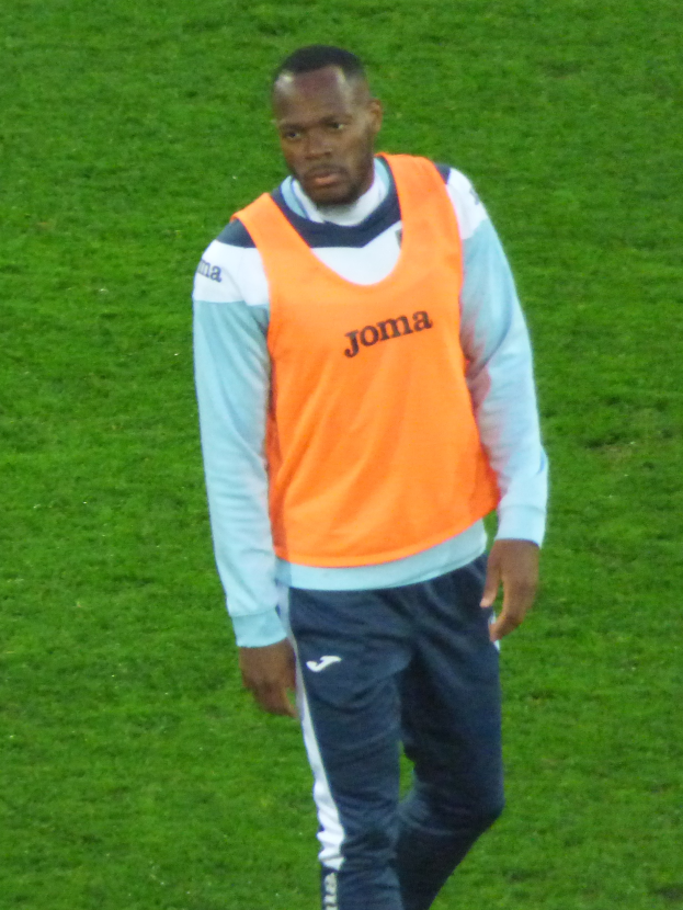Ein Mann in einem orangen T-Shirt und blauen Hosen, der scheinbar ein Fussballspieler ist, überschreitet entschlossen ein saftig grünes Feld.