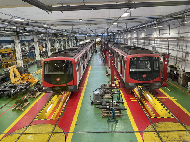Zwei Züge auf den Gleisen in einer großen Industriehalle mit Maschinen und Geräten, mit einer Wand mit Rohren und Deckenleuchten auf der rechten Seite, Teil der Daimler Group's Einrichtung.