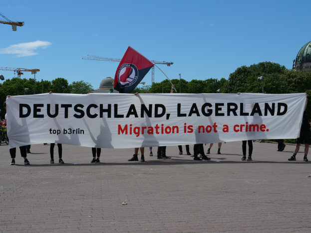 Eine Gruppe von Menschen, die draußen vor einem Gebäude mit einer Fahne und einem Banner mit der Aufschrift "Deutschland, Lagerland Migration ist kein Verbrechen" stehen.
