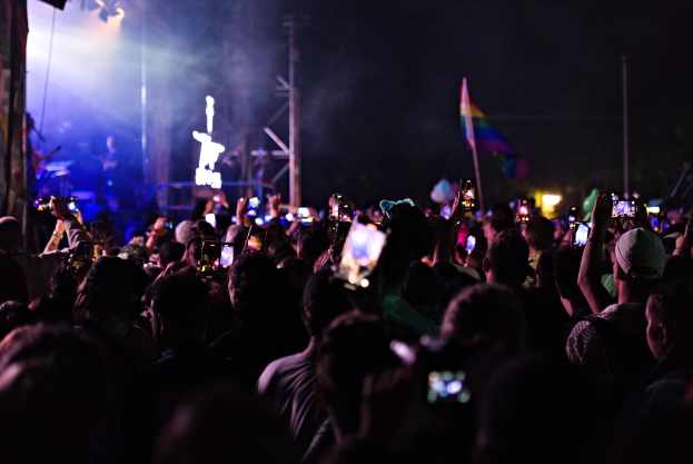 Menge bei einem Konzert mit hochgehaltenen Handys, die von Lichtern beleuchtet werden und von Pfosten und einer Fahne umgeben sind, während Musiker auf der Bühne spielen.