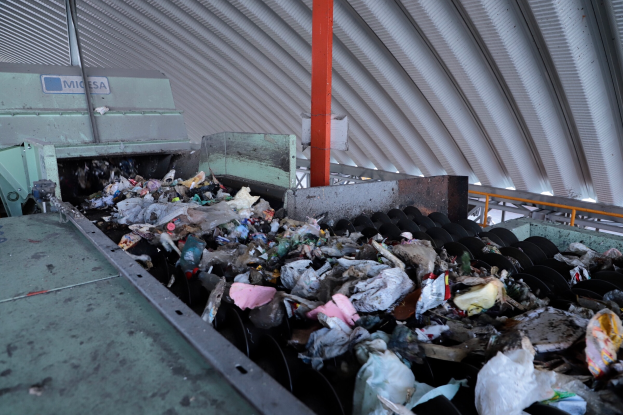 Ein großer Müllhaufen auf einem Förderband in einer Recycling-Anlage, mit Maschinen im Vordergrund und einem Metalldach oben.