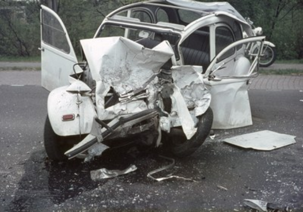 Ein schwer vorne beschädigtes Auto ist von der Straße, mit Bäumen und einem Gebäude im Hintergrund und einem weiteren Fahrzeug in der Ferne.