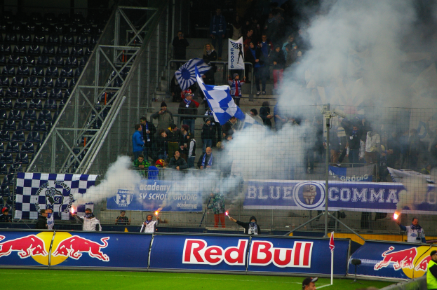 Eine Gruppe von Menschen auf einem Fußballfeld, einige halten Fahnen und Schilder, mit einem Metallzaun drum herum und Rauch, der aus dem Boden aufsteigt, und leere Stühle auf der linken Seite des Bildes.