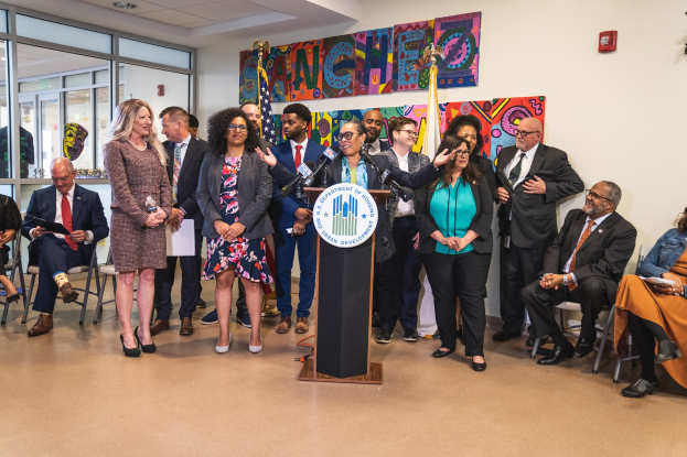 Gruppe von Menschen vor einem Rednerpult mit einem Mikrofon und einer Tafel mit der Aufschrift "Sancho-Stadtrat tritt dem Stadtrat bei", mit sitzenden und stehenden Teilnehmern, Flaggen, einem Feuerlöscher und Deckenleuchten im Hintergrund.