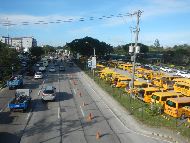 Eine belebte Straße voller Autos, Lastwagen, Bussen, Verkehrskegel, Laternenmasten, Strommasten mit Kabeln, Schildern mit Text und Maschendrahtzaun auf der rechten Seite, Bäume, Gebäude und ein bewölkter Himmel im Hintergrund.