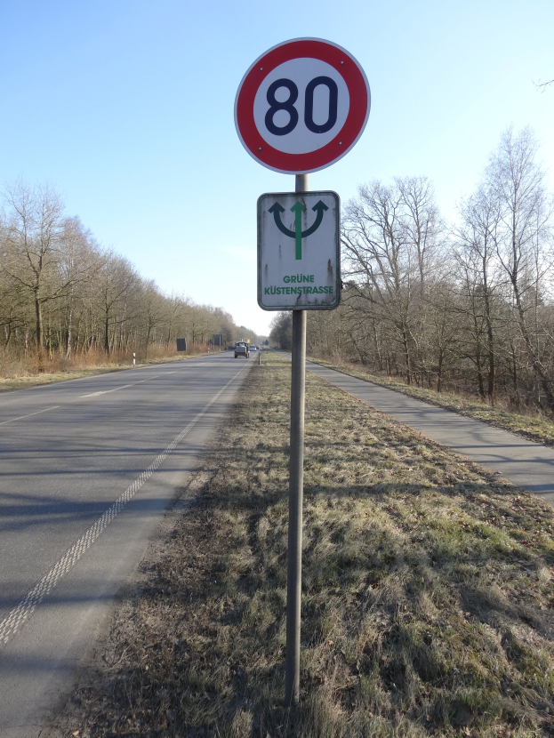 Ein Tempolimitschild am Straßenrand mit vorbeifahrenden Fahrzeugen, umgeben von Gras und Bäumen, unter einem klaren blauen Himmel.