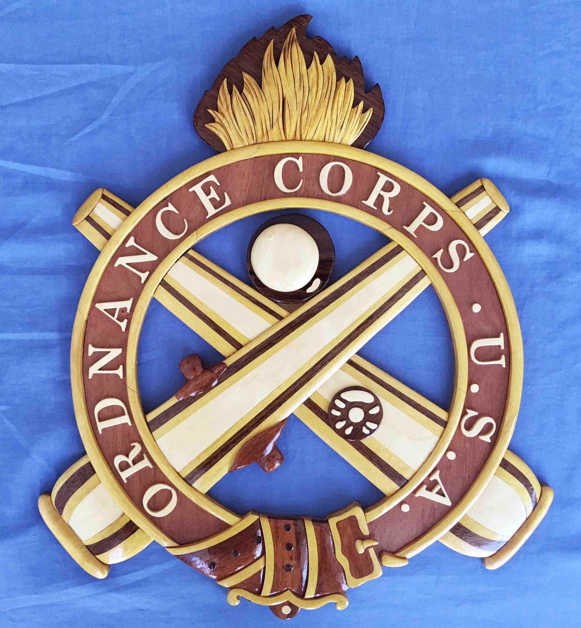 Ein hölzernes Schild mit 'Ordnance Corps' in schwarzer Schrift, das ein weißes Sternsymbol auf einem navyblauen Hintergrund mit goldener Umrandung zeigt, auf blauem Tuch montiert.