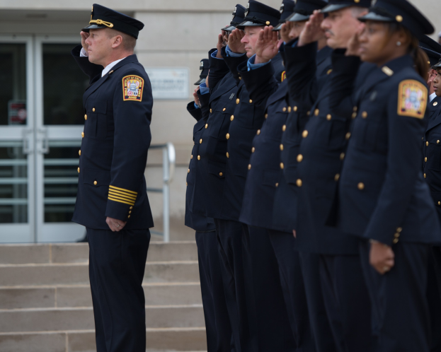 Eine Gruppe von Polizisten in Uniform und mit Mützen, die in Formation vor einem Gebäude mit Glastüren und Treppe salutieren.