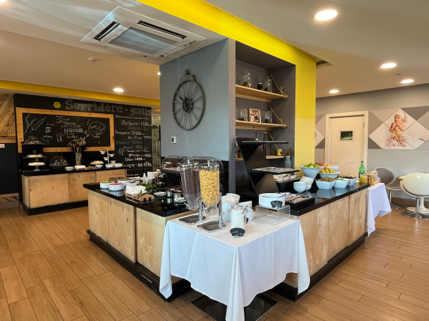 Ein Restaurant-Innenraum mit Tischen und Stühlen, Lebensmitteln, einer Tafel mit Text, einer Uhr an der Wand, Deckenleuchten, einer Klimaanlage und einer Tür im Hintergrund, das sich im Hôtel de Ville in Paris befindet.