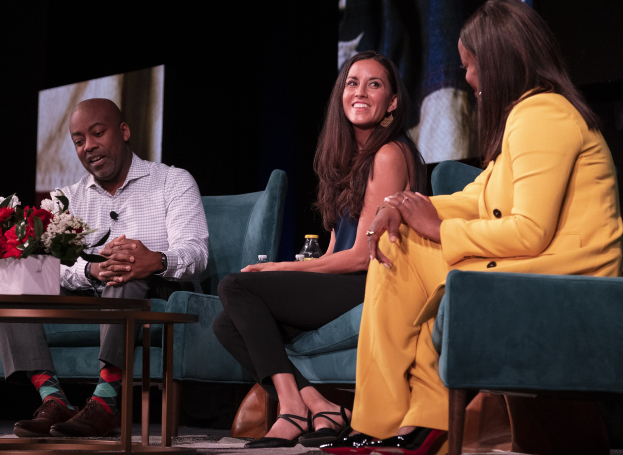 Drei Personen sitzen auf einem Sofa vor einem Bildschirm, mit einer Vase und einer Flasche auf dem Tisch dazwischen, während einer Podiumsdiskussion in einem abgedunkelten Raum.