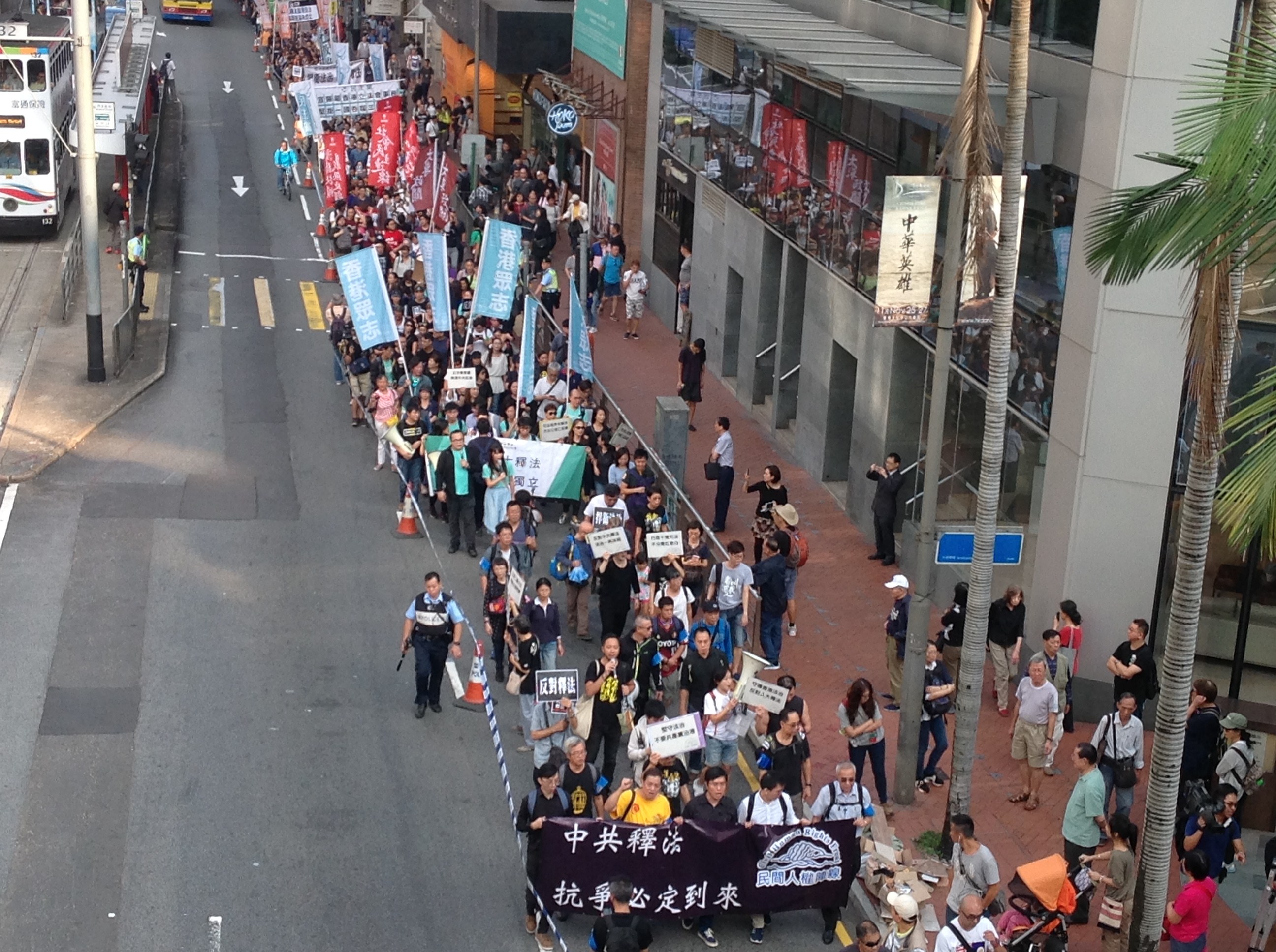 Eine große Protestmarsch in Hong Kong mit Menschen, die Schilder und Plakate tragen, die eine Straße mit Bäumen, Gebäuden, Fahrzeugen und Straßenschildern säumen.