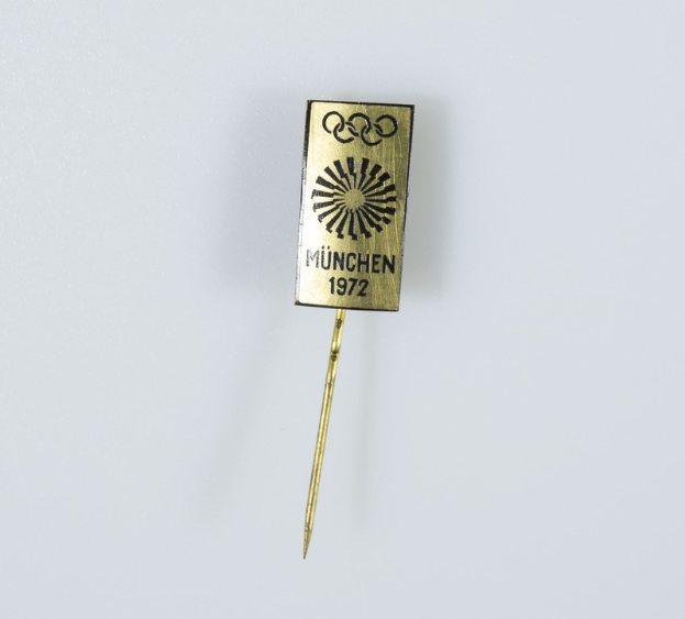 Goldene Nadel mit 'München 1972 Olympische Spiele' Text auf weißem Hintergrund.