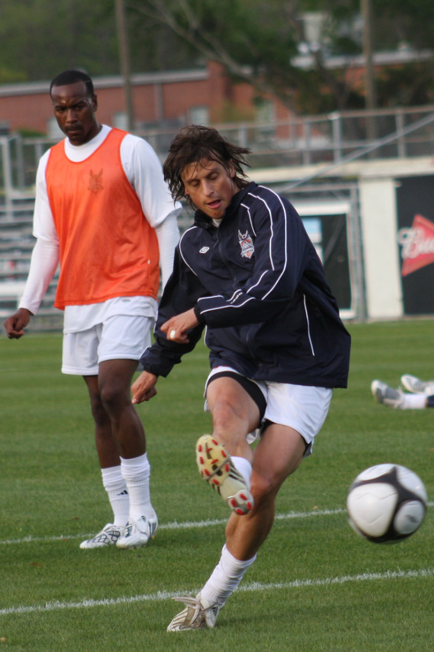 Ein Fussballspieler in einer schwarzen Jacke und weissen Shorts schiesst den Ball, mit einem anderen Spieler in einem orangen Jackett hinter ihm und einem Zaun mit Bäumen im Hintergrund.