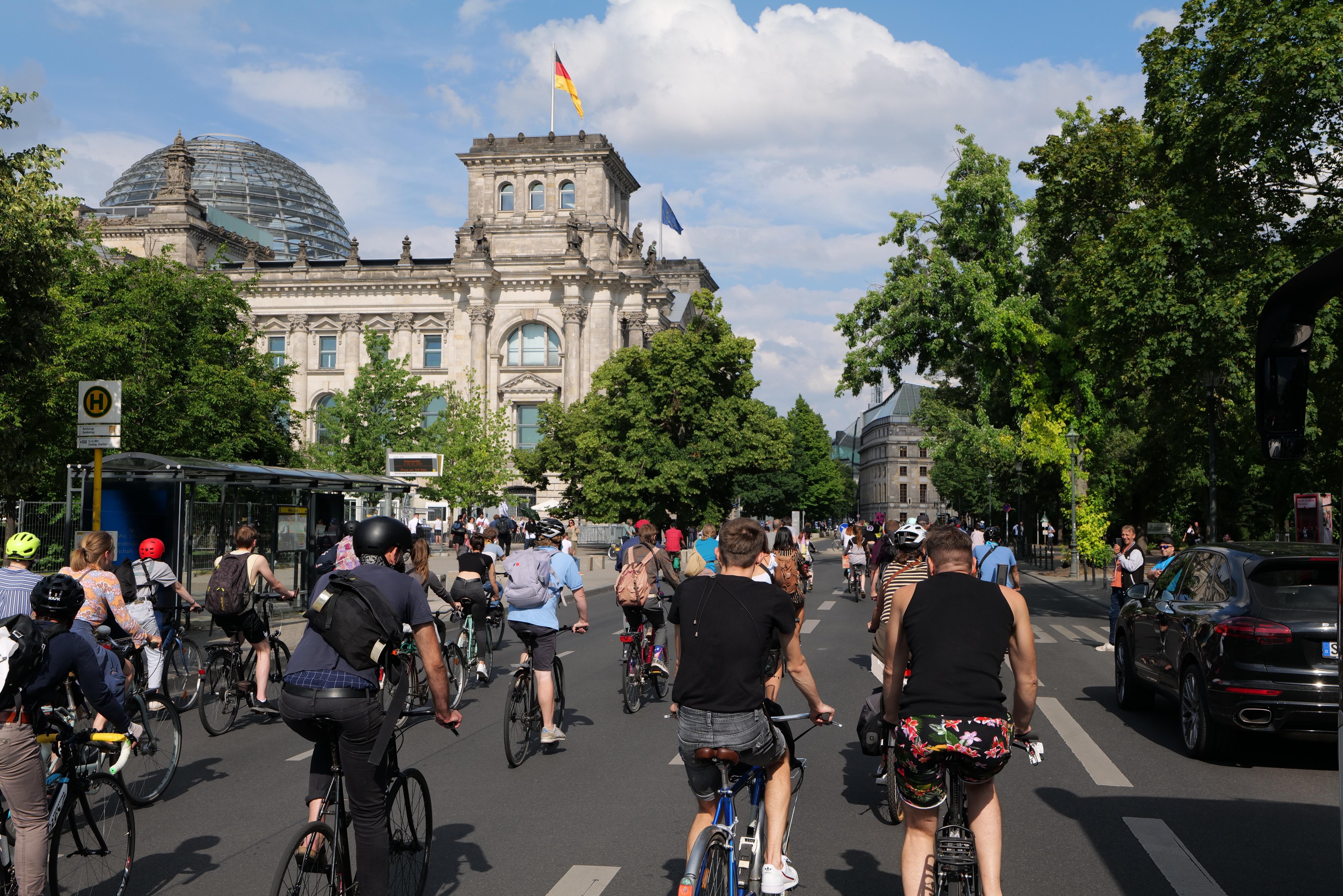 Eine Gruppe von Menschen, die Fahrräder auf einer von Bäumen gesäumten Straße in Berlin, Deutschland, fährt, mit Gebäuden auf der linken Seite und einer Bushaltestelle auf der rechten Seite unter einem bewölkten Himmel, eine Flagge sichtbar auf einem der Gebäude.
