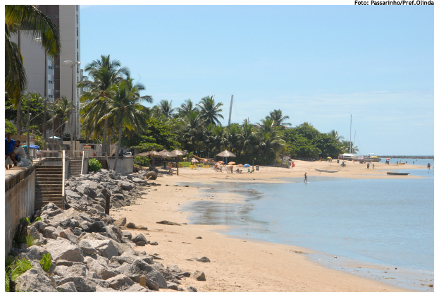 Eine Strandszene mit Wasser auf der rechten Seite, Felsen und einem Gebäude auf der linken Seite, Bäumen im Hintergrund, Sand im Vordergrund und vielen Menschen verteilt.