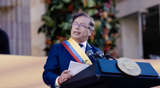Ein Mann in einem blauen Anzug und Krawatte steht an einem Podium, hält Papiere und spricht in ein Mikrofon, mit einer Pflanze und einer Wand dahinter, was eine formelle Umgebung suggeriert.