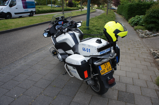 Ein Polizei-Motorrad am Straßenrand mit einem Helm darauf, umgeben von Bäumen, Masten, Gebäuden, Pflanzen, Gras, Steinen und anderen Fahrzeugen.