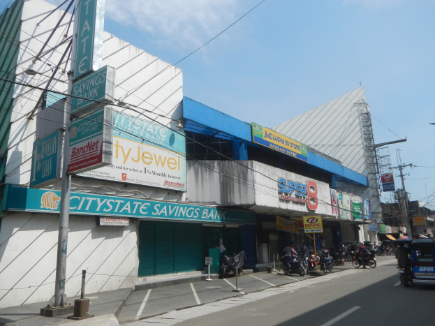 Eine belebte Stadtstraße mit Fahrzeugen, Fußgängern, Strommasten, Gebäuden und der Citystate Sparbank in Cebu City im Hintergrund bei bewölktem Himmel.