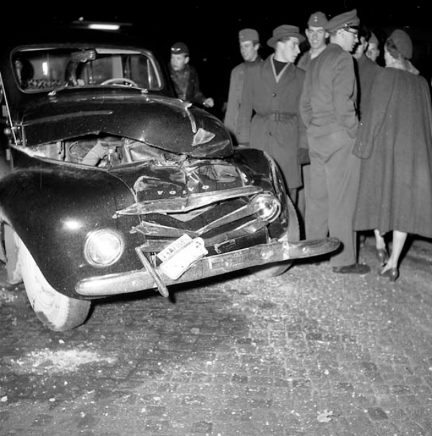 Ein Schwarz-Weiß-Bild eines beschädigten Autos am Straßenrand, umgeben von einer Gruppe von Menschen, die besorgt auf das Auto schauen.