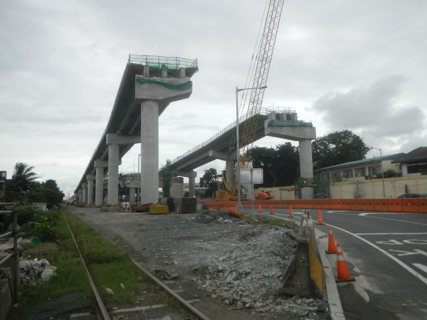 Eine Baustelle mit einer Brücke im Hintergrund, eine Straße mit Absperrkegeln auf der rechten Seite, Steine und Gras auf dem Boden, eine Eisenbahnschiene auf der linken Seite, Bäume und Gebäude auf beiden Seiten der Straße und ein bewölkter Himmel.