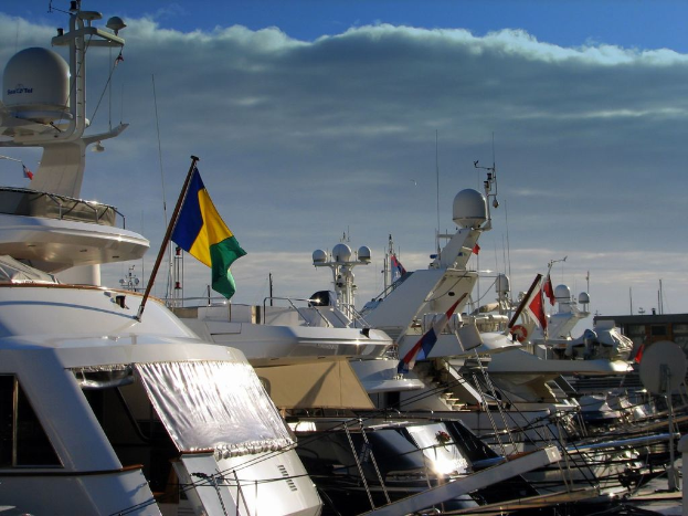 Ein weißes Boot mit einer Flagge ist auf einem Schiffbauplatz mit anderen Booten geparkt, unter einem blauen Himmel mit Wolken.