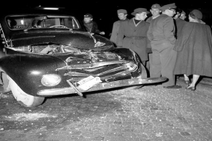 Eine Schwarz-Weiss-Szene eines beschädigten Autos am Straßenrand, umgeben von einer Gruppe besorgter Menschen.