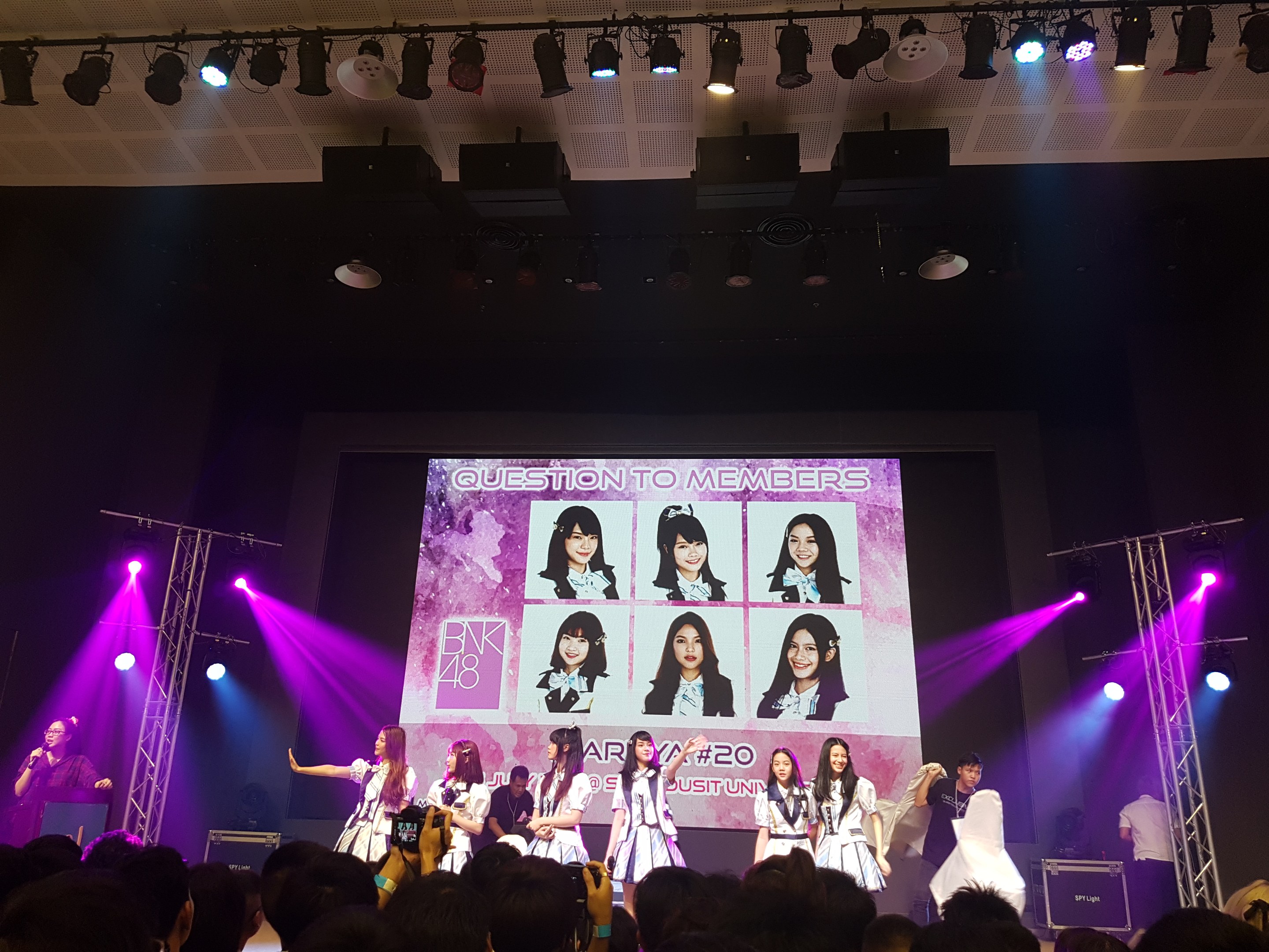 Eine Gruppe von Menschen performt auf einer hell beleuchteten Bühne mit Trägern, mit einem Sänger mit Mikrofon links und einem Musiker, der ein Instrument spielt, während eine Menge mit Kameras unten und ein großer Bildschirm Bilder der K-Pop-Gruppe BK48 zeigt.