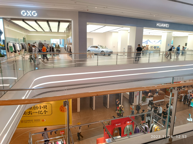 Das Innere eines Einkaufszentrums mit Menschen, die herumlaufen, Glasgeländern, Kleider auf Bügeln, Texttafeln, Säulen, Deckenlampen und einem geparkten Auto im Hintergrund, mit einem GXG- und Huawei-Partnerschafts-Wasserzeichen unten.